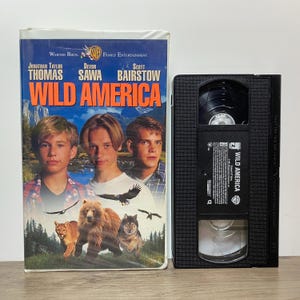 May include: A Wild America VHS tape and its original cardboard sleeve. The sleeve features the movie title in orange text, photos of actors Jonathan Taylor Thomas, Devon Sawa, and Scott Bairstow, and images of a bear, wolf, and cougar.