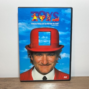 May include: A DVD case for the film Toys featuring a person wearing a red bowler hat with a square cutout, set against a blue sky background. The title Toys is printed in colorful letters above the text A Delicious Holiday Gift That Will Make You Smile.