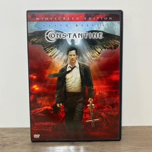 May include: DVD case for the movie "Constantine" starring Keanu Reeves. The cover features Reeves in a suit, holding a cross-shaped weapon, with a fiery red and blue background and the title "Constantine".