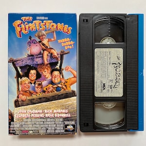 May include: VHS video cassette tape of the 1994 film The Flintstones. The cover features a cartoon image of the Flintstones family riding in a car. The text on the cover reads "The Flintstones" and "Yabba-Dabba-Doo!" The actors John Goodman, Rick Moranis, Elizabeth Perkins, and Rosie O'Donnell are listed below the image.