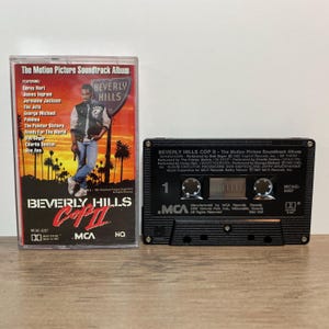 May include: A vintage cassette tape and case for the "Beverly Hills Cop II" soundtrack album. The case features a movie poster design with the film's title and artist names. The black cassette tape is labeled with the album's title and MCA Records.