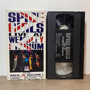 May include: A VHS tape and its case featuring the Spice Girls. The case has the text "SPICE GIRLS LIVE AT WEMBLEY STADIUM" in red, white, and blue. The tape is black with white text, including the band's name and the venue.