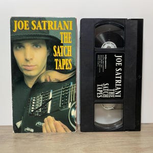 May include: A Joe Satriani The Satch Tapes VHS cassette and its cardboard slipcase. The cover features a portrait of Joe Satriani wearing a hat and holding an electric guitar. The black plastic tape has a label with the title and copyright info.
