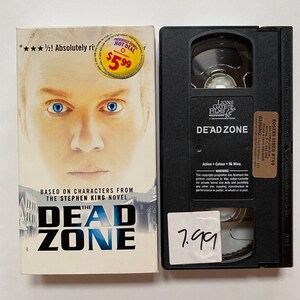 May include: A VHS tape and book of "The Dead Zone." The book cover features a man with blue eyes and the title in bold letters. The VHS tape is black with the title "DE'AD ZONE" and a sticker with the number 7.99.