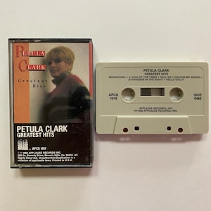 Peut inclure: Une cassette audio blanche avec une étiquette noire. L'étiquette porte le texte "Petula Clark Greatest Hits" et les titres des chansons "Downtown", "A Sign of the Times", "Call Me", "Colour My World", "Strangers in the Night", et "Hello Dolly". La cassette est étiquetée "APCS 1015" et "Face A".