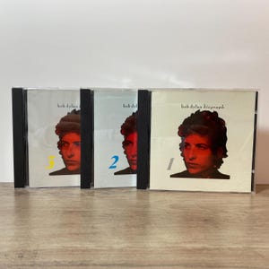 May include: Three Bob Dylan CD albums in jewel cases. The album covers feature a portrait of Bob Dylan with the text "bob dylan" and "bob dylan biograph". The CDs are numbered 1, 2, and 3.