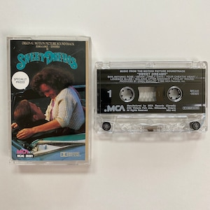 Peut inclure: Une cassette audio de la bande originale du film "Sweet Dreams" avec Jessica Lange et Ed Harris. La cassette est en plastique transparent avec une étiquette noire. L'étiquette porte le titre du film, les noms des acteurs et le logo MCA Records. La cassette est dans un boîtier en plastique transparent avec un fond blanc. Le boîtier porte le titre du film, les noms des acteurs et le logo MCA Records. Le boîtier comporte également un autocollant indiquant "Prix Spécial".
