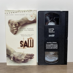 May include: A Saw film VHS tape and its cardboard slipcase. The white slipcase features a graphic of a foot and a hand, the title Saw, and the text Every puzzle has its pieces. The black plastic VHS cassette has the Lions Gate Home Entertainment logo.