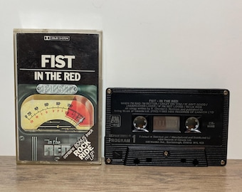 May include: A black cassette tape and case with the text "FIST IN THE RED". The cassette case has a graphic of a radio dial. The cassette tape has the text "PROGRAM 1" and song titles.