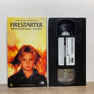 May include: A VHS tape of the film Firestarter featuring a yellow cover with a young person surrounded by flames and the text Charlie McGee is Stephen King's FIRESTARTER Will she have the power...to survive? The black cassette has a white label.