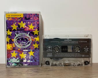 May include: A clear cassette tape of the U2 album "Zooropa" with a purple, yellow, and white cover. The cassette case is transparent, and the tape inside is visible. The album title and tracklist are printed on the cassette.
