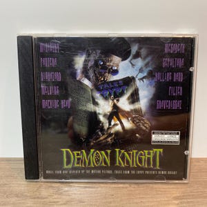 May include: A CD case for the movie "Demon Knight." The cover features a ghoulish figure with glasses holding a book titled "Tales from the Crypt." The CD case lists the bands Ministry, Pantera, Biohazard, Melvins, Machine Head, Megadeth, Sepultura, Rollins Band, Filter, and Gravediggaz.