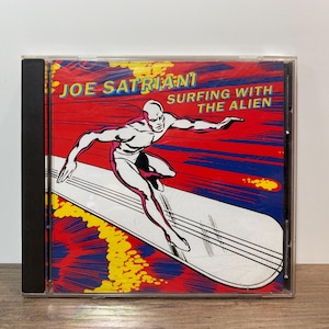 May include: A Joe Satriani CD case featuring the album "Surfing with the Alien." The cover art depicts a silver-skinned figure surfing on a silver surfboard against a red, blue, and yellow background. The artist's name is at the top.