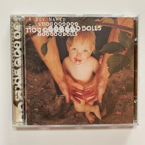 May include: A CD case with a photo of a baby with a messy face, sitting in the hands of an adult. The CD case has the title "A Boy Named Goo" and the band name "Goo Goo Dolls" printed on the front.