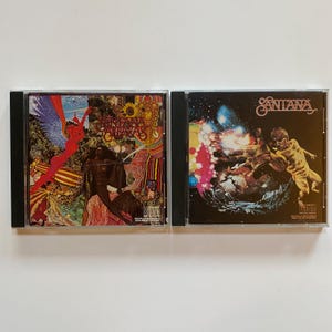May include: Two compact discs, both featuring the band Santana. The first disc is "Santana Abraxas" with a psychedelic cover featuring a woman in a red dress and a bird. The second disc is "Santana" with a black and white cover featuring a man in a white suit flying through the air.