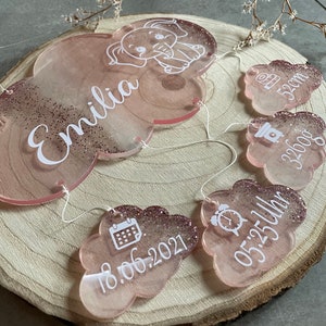 Könnte beinhalten: Rosa und glitzernde wolkenförmige Acrylschmuckstücke mit weißem Text und einem Elefanten-Design. Die Ornamente enthalten den Namen "Emilia", Maße, Datum und Uhrzeit. Die Ornamente sind mit weißer Schnur verbunden.