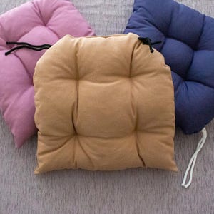 Könnte beinhalten: Drei quadratische Sitzkissen in Rosa, Beige und Marineblau. Jedes Kissen verfügt über ein gestepptes Design mit vier Vertiefungen und an den Ecken befestigten weißen oder schwarzen Stoffbändern zur Befestigung an einem Stuhl oder Möbelstück.