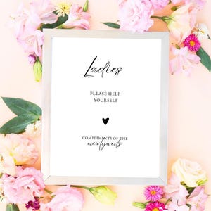 May include: A white sign with black text that reads "Ladies, Please Help Yourself, Compliments of the Newlyweds". The sign is framed in a silver frame and surrounded by pink flowers.