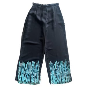 Puede incluir: Pantalones negros de pierna ancha con cintura alta y abertura en el dobladillo. La parte inferior de los pantalones presenta un diseño abstracto en azul claro. Los pantalones están hechos de un material brillante y sedoso.