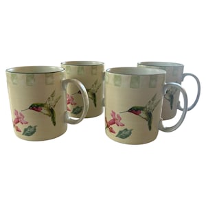 May include: Set of four ceramic mugs with a cream-colored base. Each mug features a green and red hummingbird design with pink flowers and green leaves. The rims have a green square pattern. The mugs have silver-colored handles.