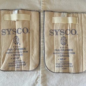 May include: Two beige canvas pan grabbers with dark blue text reading SYSCO, 8 1/2 x 11 PAN GRABBER, PROTECTS HANDS FROM GREASE HEAT, DISTRIBUTED BY SYSCO CORPORATION HOUSTON, TEXAS 77077 #4799813. Each features a horizontal cream-colored fabric strap.