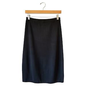 Puede incluir: Una falda negra colgada de una percha de madera. La falda es de color negro liso y parece estar hecha de una tela suave. La falda llega a la rodilla.