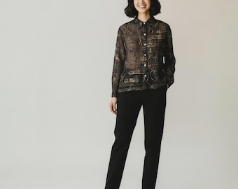 May include: A person wearing a sheer, button-down blouse with a dark, ornate pattern, paired with black trousers and gold heels. The blouse features a detailed design in shades of gold and black.
