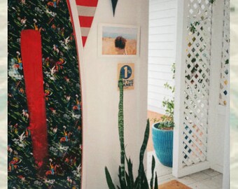May include: A black and white patterned surfboard with a red stripe down the center. The surfboard is leaning against a white wall with a blue and white pennant that says "NEWPORT". A potted snake plant is in the foreground.