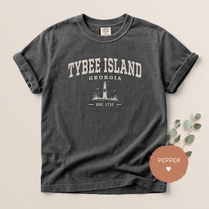 May include: Dark gray t-shirt with rolled sleeves. The front features the text "TYBEE ISLAND GEORGIA" above a lighthouse illustration and "EST. 1733". The shirt has a vintage, worn look.