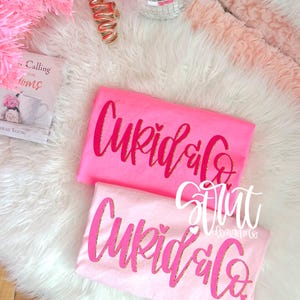 May include: Two pink t-shirts with the text "Cupid & Co" in a cursive font with gold glitter accents. One shirt is a brighter pink, the other is a lighter pink. The shirts are on a white fluffy surface.