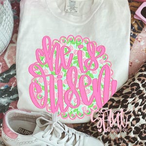 May include: White t-shirt with a pink floral design that reads "She is Risen." The design is surrounded by a pink scalloped border. The shirt is paired with white sneakers with pink accents and a leopard print fabric.