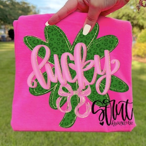 May include: A folded pink t-shirt featuring a green four-leaf clover with a leopard print pattern. The word "Lucky" is written in pink script, with a pink bow below. The text "Strut Design & Co" is in black script.