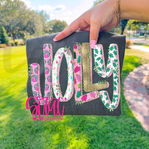 May include: Dark gray t-shirt with the word "JOLLY" in a festive design. The letters are decorated with pink, gold, and green patterns, including Christmas trees and holly. The shirt also features a pink logo at the bottom.