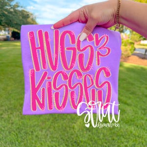 May include: A lilac-colored t-shirt with the words "HUGS KISSES" in a bright pink, outlined in yellow. The shirt also features the text "stunt" and "vloesign&co" in white script. The shirt is held up against a background of green grass and a blue sky.