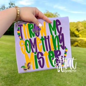 May include: A light purple t-shirt with the text "THROW ME SOMETHING MISTER" in green, yellow, and purple. The shirt features Mardi Gras-themed embellishments, including a fleur-de-lis and a mask. The shirt is held up by a person.
