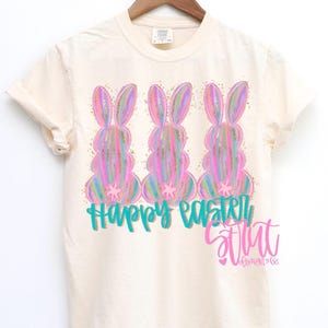 May include: Cream-colored t-shirt featuring three colorful Easter bunny designs with gold glitter accents. The words "Happy Easter" are written in teal, with a pink script logo below.