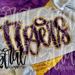 May include: A gray long-sleeved shirt with the word "Tigers" in a large, decorative font. The letters are black with a purple and gold pattern, outlined in gold. The shirt also features the text "Starr Designs & Co."