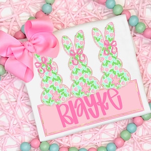 May include: White t-shirt featuring three bunny silhouettes with floral patterns in pink and green, each adorned with a pink bow. The name "Blayke" is displayed in pink script on a pink rectangle with a gold border.