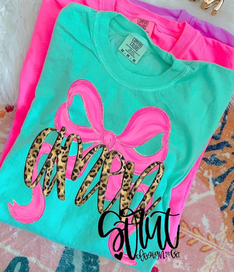 Puede incluir: Una camiseta turquesa con un gran lazo rosa y la palabra "Girl" en una fuente de estampado de leopardo. La palabra "Strut" est&aacute; escrita en gui&oacute;n negro debajo del lazo. Se ven otras dos camisetas en rosa y morado en el fondo.