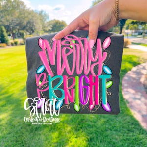 May include: Dark gray t-shirt with the words "MERRY BRIGHT" in pink, gold, and teal. The design includes colorful Christmas lights. The shirt is held up against a green grassy background.