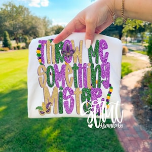 Op de afbeelding: Wit t-shirt met de tekst "THROW ME SOMETHING MISTER" in goud, paars en groene glitter. Het ontwerp bevat een fleur-de-lis en een masker, met Mardi Gras kralen. Het shirt wordt door een persoon omhoog gehouden.