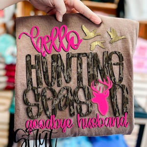 May include: A brown t-shirt with the words "Hello Hunting Season goodbye husband" in pink script. The words "HUNTING SEASON" are in camouflage print. Gold-colored flying ducks and a pink deer head are also featured.