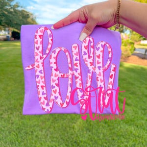 May include: A lilac-colored t-shirt with the words "Love You" in a pink, heart-filled design. The shirt is held up against a backdrop of green grass and a blue sky.
