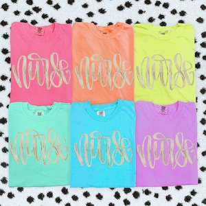 May include: Six short-sleeved crew neck t-shirts in bright neon colors including pink, orange, yellow, mint green, blue, and purple. Each shirt features the word Nurse printed across the chest in a large, gold-colored cursive script font.