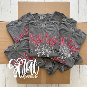 May include: A collection of charcoal gray t-shirts featuring a red script graphic that reads Sic 'em above a white line-art illustration of a bulldog face, with the text EST 1785 and Athens GA printed in black.