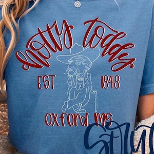Op de afbeelding: Een blauw T-shirt met ronde hals met de rode tekst Hotty Toddy, de witte tekst EST 1848 en de rode tekst OXFORD, MS. Een witte lijntekening van een man in een pak en cowboyhoed die een wandelstok vasthoudt, staat centraal tussen de tekst.