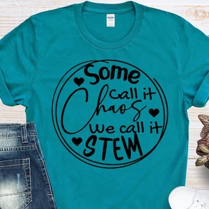 May include: Teal t-shirt with the text "Some call it chaos, we call it STEM" in a black circular design. The shirt is paired with blue jeans and white sneakers. A small potted plant is also in the image.