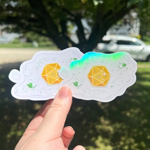 May include: Two white stickers with a cloud-like shape, each featuring a yellow twenty-sided die with the number 20 visible. Green leaf and dot details are scattered around the die. One sticker has a holographic effect.