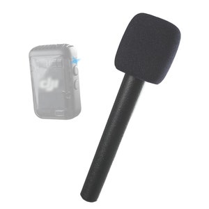 Könnte beinhalten: Ein schwarzes Mikrofon mit Schaumstoff-Windschutz und schwarzem Griff. Das Mikrofon ist für die Aufnahme von Audio konzipiert. Im Hintergrund befindet sich eine transparente Hülle für ein kleines elektronisches Gerät.