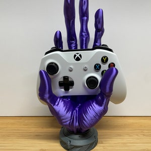 May include: A purple 3D printed hand holding a white Xbox controller. The hand is on a gray base.